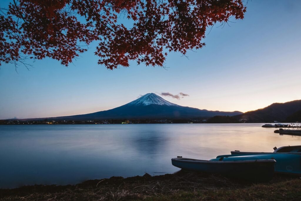 Mount Fuji at sunset, viewed across a calm lake with autumn leaves and boats in the foreground.