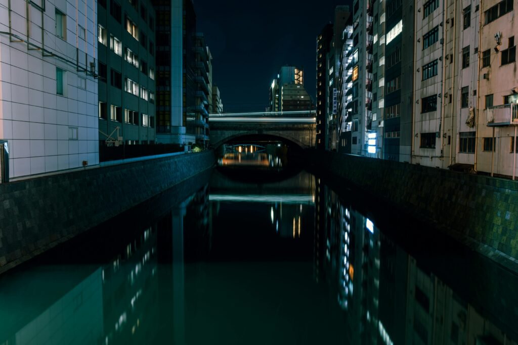 Night view of a Tokyo canal with tall buildings and their lights reflected in the water. A bridge connects the buildings across the canal.