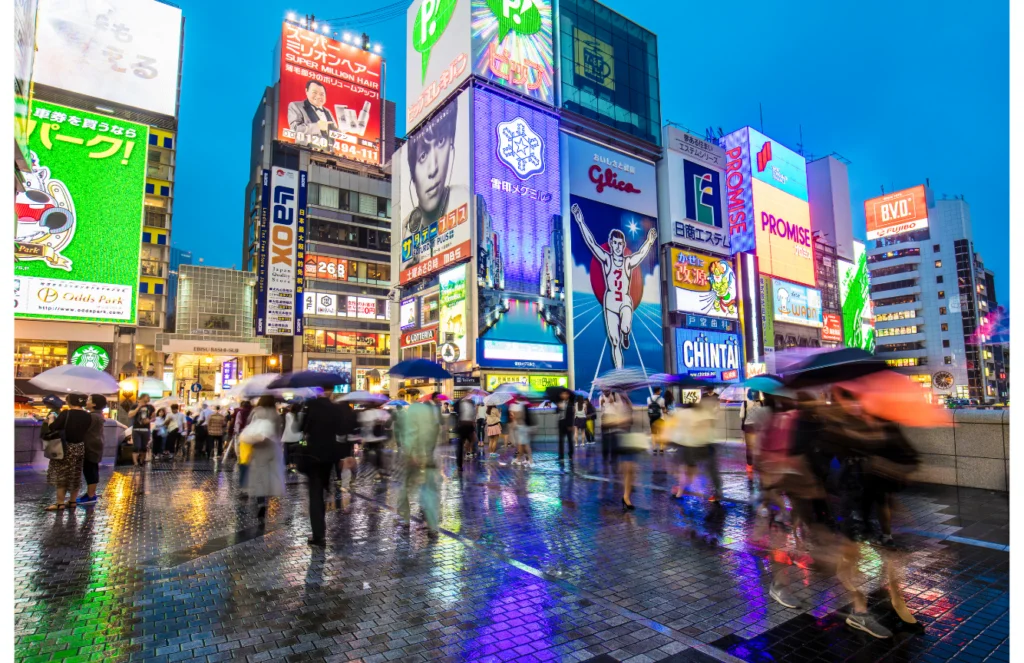 Crowds of people with umbrellas walk through Dotonbori in Osaka during the evening, with colorful neon signs reflecting off the wet pavement. The iconic Glico running man sign is brightly lit in the center, surrounded by other vibrant advertisements and billboards.